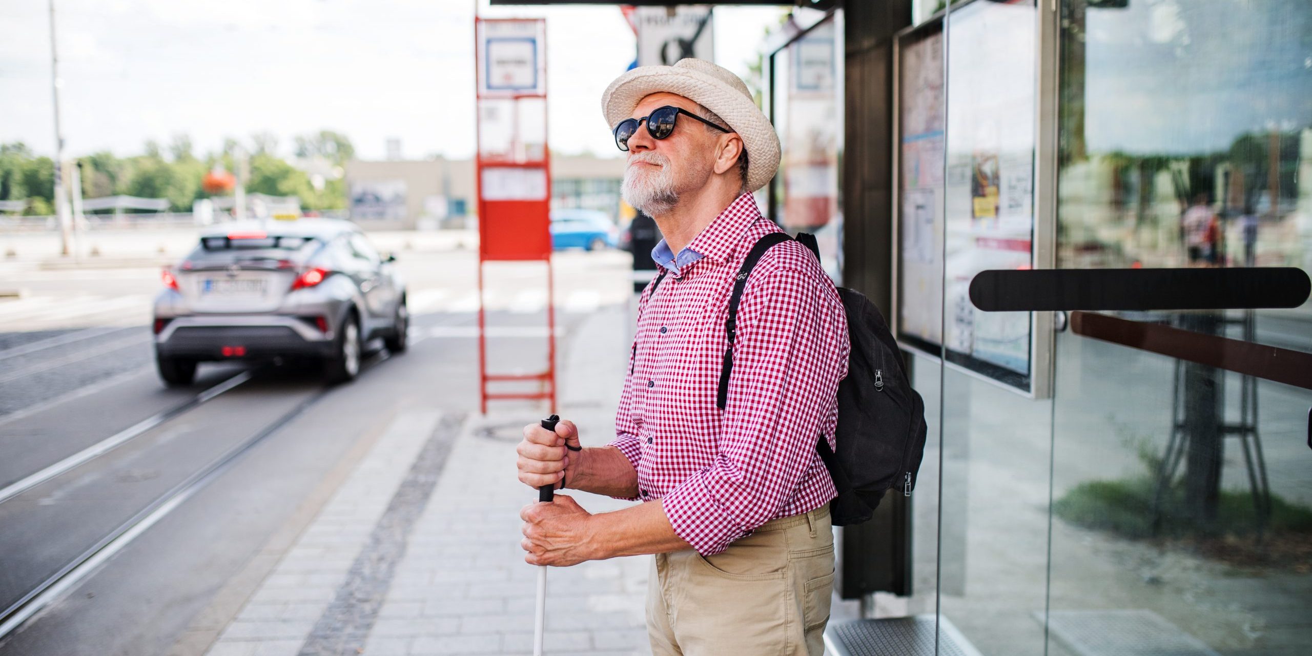 A senior blind man with white cane waiting at bus stop in city.