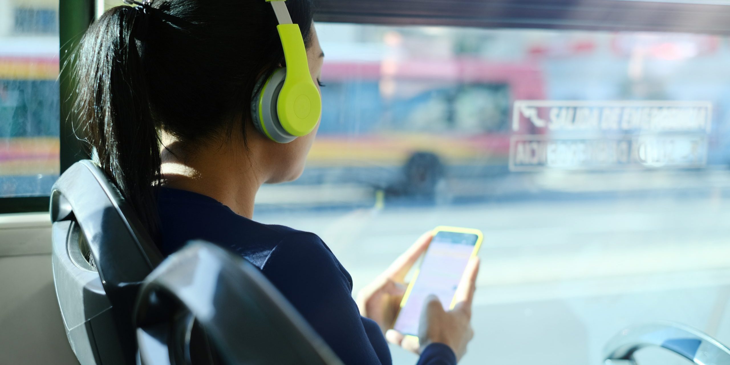 Woman With Headphones Listening To Music While Commuting To Work Using Public Transportation. Latin American Woman Sitting In A Bus And Using Mobile Phone.
